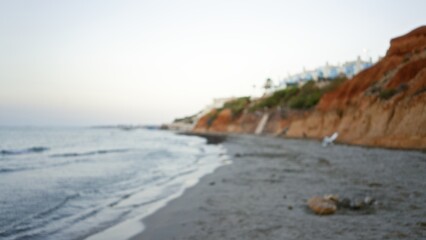 Blurry view of a serene outdoor beach with sandy shores, calm ocean, and gently sloping coastal cliffs at sunrise creating a peaceful atmosphere