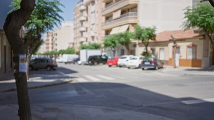 Blurred city street scene with cars parked on the roadside, trees lining the pavement, residential buildings in the background, and a poster on a tree trunk, providing a dreamy, urban atmosphere.