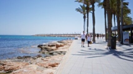 Blurred background of an outdoor seafront promenade with palm trees, a walkway, and a defocused couple walking by the sea on a sunny day