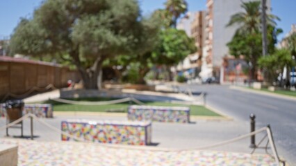 Obraz premium Blurred background of an outdoor public street with colorful benches, trees, and buildings, palm trees lining the road under a bright sunny sky