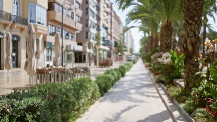 Fototapeta premium Blurry outdoor pedestrian walkway with defocused buildings, palm trees, and greenery lining the path on a sunny day.