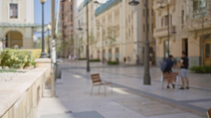 Blurred people on outdoor urban street with benches, defocused buildings, and bokeh effect creating a soft, out of focus background in a sunny cityscape.