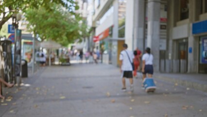 Blurred people walking in an outdoor shopping area with trees and storefronts in the background, showcasing a typical urban street scene with men and women, defocused for a bokeh effect