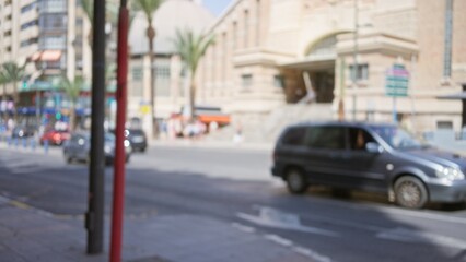 Outdoor city street scene with blurred background, featuring cars, buildings, sidewalks, and some people in the distance in a bokeh effect