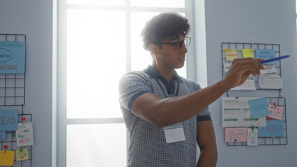 Young hispanic man in an office pointing at something with a pen against a backdrop of notes and charts on the wall under daylight.