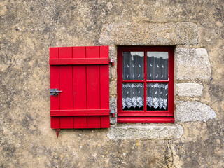 A rustic stone wall in Brittany with a red wooden window and shutter, featuring lace curtains.