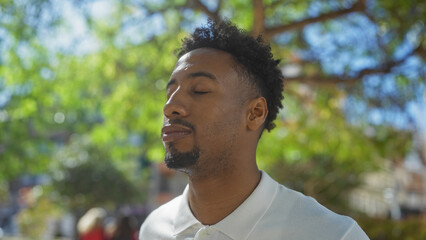 Handsome young african american man with a beard enjoying a sunny day outdoors in an urban park with closed eyes, surrounded by vibrant greenery.