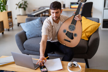 Arab man combines playing an oud with jotting down notes in a warm, inviting living room, surrounded by personal items and a colorful interior that reflect a creative and relaxed atmosphere.