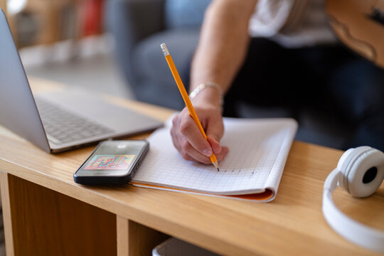A musician takes notes on paper with a pencil while holding an oud, with a laptop and other musical elements around, indicating a creative process unfolding in a comfortable and homey environment.