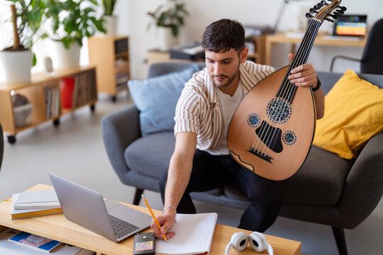 Arab male musician reaches for a pencil on a table with a notebook and an oud beside it, symbolizing a moment of creativity and musical reflection in a peaceful and artistic home environment.