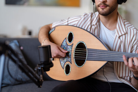 Close-up of a man wearing headphones, strumming an oud while recording in a cozy living room, capturing the essence of home music production and passion for musical creation.