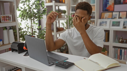 Young man in a home decor store with a laptop holding glasses and rubbing his eyes