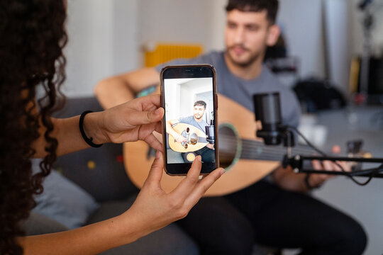 woman captures a video of a musician playing an oud on her smartphone, showcasing a casual music recording session in a relaxed indoor setting with a focus on content creation and musical performance