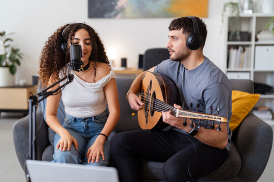 A young man plays an oud while a woman sings into a microphone, both wearing headphones, engaging in a focused recording session in a cozy home setting emphasizing creativity and teamwork.