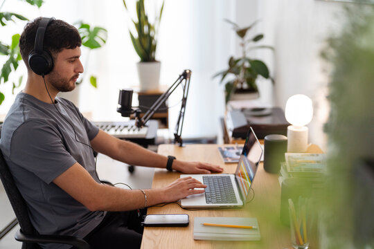 Arab young man wearing headphones works on a laptop in a home studio with audio editing software, showcasing a focused setup perfect for music production in a modern, indoor workspace.