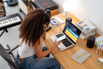 A young woman with headphones uses a smartphone while editing audio on a laptop in a home studio, capturing the multitasking nature of modern music production in a cozy environment.