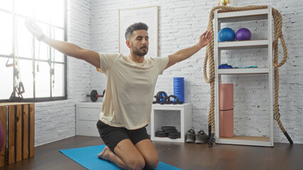 A handsome young hispanic man performs exercises on a mat in a gym, surrounded by various sports...