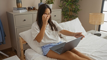 Fototapeta premium Young woman with brunette hair sitting on a bed in a bedroom adjusting her glasses while using a laptop, surrounded by home decor items.