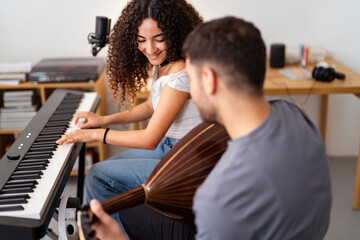 A young woman plays the keyboard with a microphone set up beside her, while a man plays an oud, focusing intently on his instrument, with recording equipment and cozy home decor in the background.