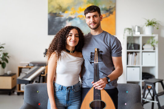 man holds an oud as he stands beside a smiling woman, both posing in a warmly decorated room with various musical instruments and shelves in the background, creating a relaxed and inviting atmosphere.