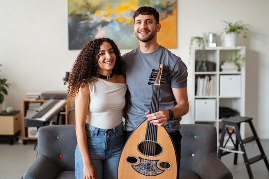 man holds an oud while standing beside a smiling woman in a contemporary living room, with a colorful painting and musical equipment creating a vibrant and friendly setting for creativity.