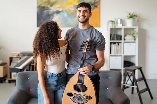 man holds an oud while standing beside a smiling woman in a contemporary living room, with a colorful painting and musical equipment creating a vibrant and friendly setting for creativity.