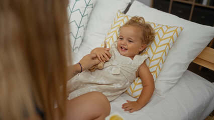 Blonde toddler girl smiling on a bed in a cozy bedroom while playing with an adult woman, capturing a warm childhood moment indoors.