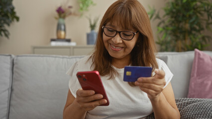 Hispanic woman using a smartphone and holding a credit card in her living room, surrounded by comfortable home decor and greenery, showcasing online shopping.