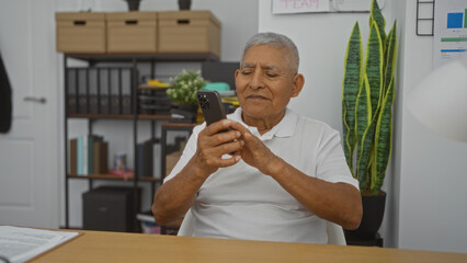 An elderly hispanic man with grey hair is using a smartphone in an office setting with shelves, folders, and a plant in the background.