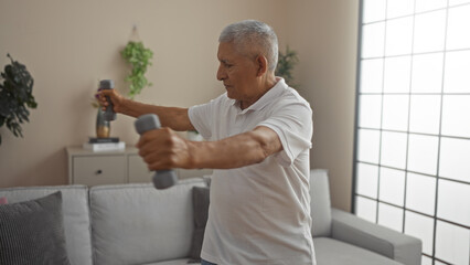 Fototapeta premium A grey-haired hispanic man exercises with dumbbells in a cozy living room, demonstrating health and fitness at home in a warm, well-lit interior setting.