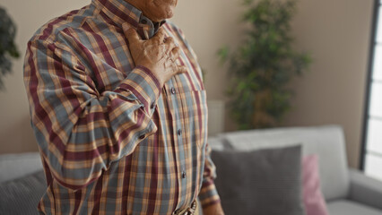 Senior man in plaid shirt indoors with hand on chest, indicating heart pain, in cozy living room setting.