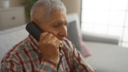 Middle-aged hispanic man with grey hair talking on a landline phone in a cozy living room.