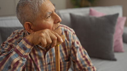 A mature hispanic man with grey hair and a patterned shirt rests on a wooden cane in a cozy living room.