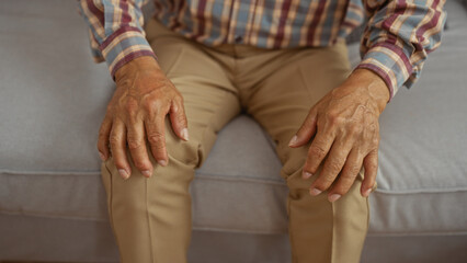 Middle-aged man with hands resting on knees sitting indoors on a sofa in a living room wearing a plaid shirt and khaki pants.