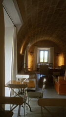 Cozy, warmly-lit cafeteria with rustic decor featuring white curtains and arched ceilings, a wooden table with chairs, and a large window letting in natural light