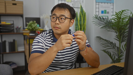 A young asian man sitting in a modern office, wearing glasses and a striped shirt, looks thoughtful while holding a pen.