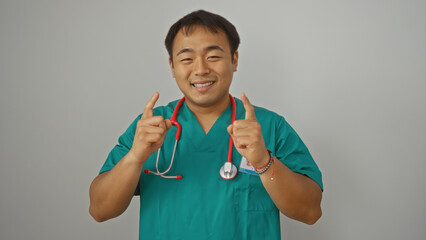 A young handsome chinese man in medical scrubs with a stethoscope smiles and points up, isolated against a white background.