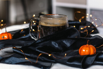 Black coffee in a glass mug on a table with a black tablecloth and orange pumpkins with a face. Halloween Table Decoration