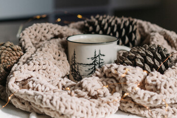 Coffee with milk in a white mug in a warm knitted blanket and big cones next to it. Cozy Christmas. Happy Holidays. Space for text