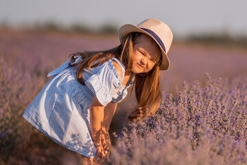 Little girl in in white and blue dress across field of purple lavender smelling lavender at sunset.