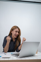 A woman is sitting at a desk with a laptop open, smiling and celebrating