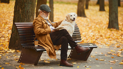 Mature man sitting on bench with his beloved, cute pet, Retriever dog and reading book in autumn...