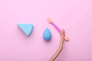 Doll's hands holding makeup accessories on pink background
