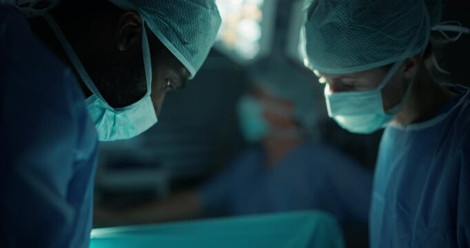 Diverse Surgeons and Nurses Team Dressed in Full Protective Gear, Talk and Coordinate Around the Operating Table, Saving Life of a Patient with Their Combined Skills and Medical Training