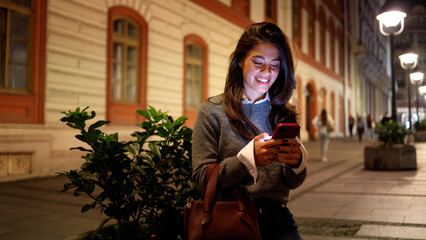 Happy woman texting on smartphone while taking a walk downtown in the evening