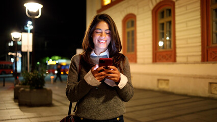 Happy woman walking through the brightly lit city streets and using phone