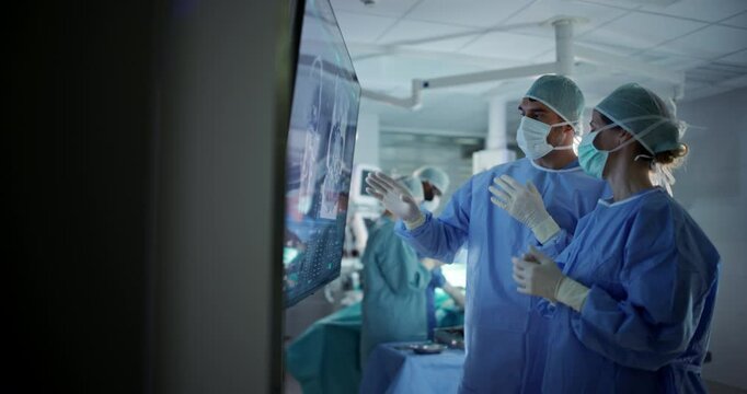 Portrait of a Young Female and Male Surgeons Reviewing Patient's Scans on a Large Screen in a Dimly Lit Operating Room. Their Team of Assistants and Nurses Preparing for a Life-Saving Procedure