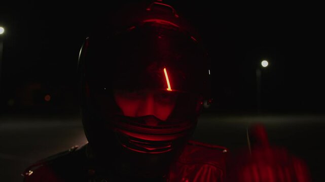 Young handsome man in leather gear closes toned visor on motorcycle helmet and lights of night city and letters of neon advertising are reflected in it. Slow motion. Passion for extreme racing