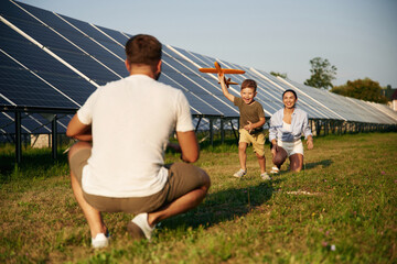 Running to a parent with toy plane. Father, mother and little son are outdoors near solar panels