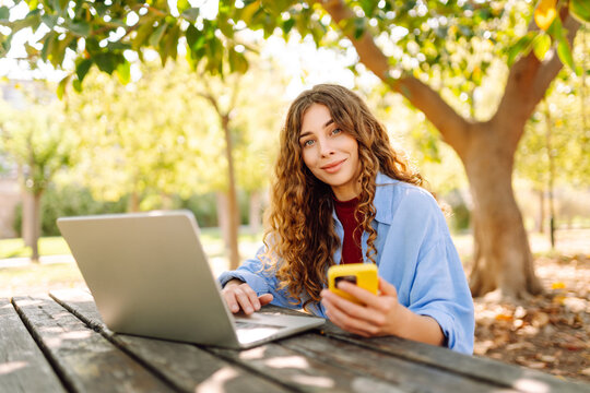 Young smiling student sit on bench in spring park outdoors, rest, use laptop, mobile cell phone browsing internet. People urban lifestyle concept.